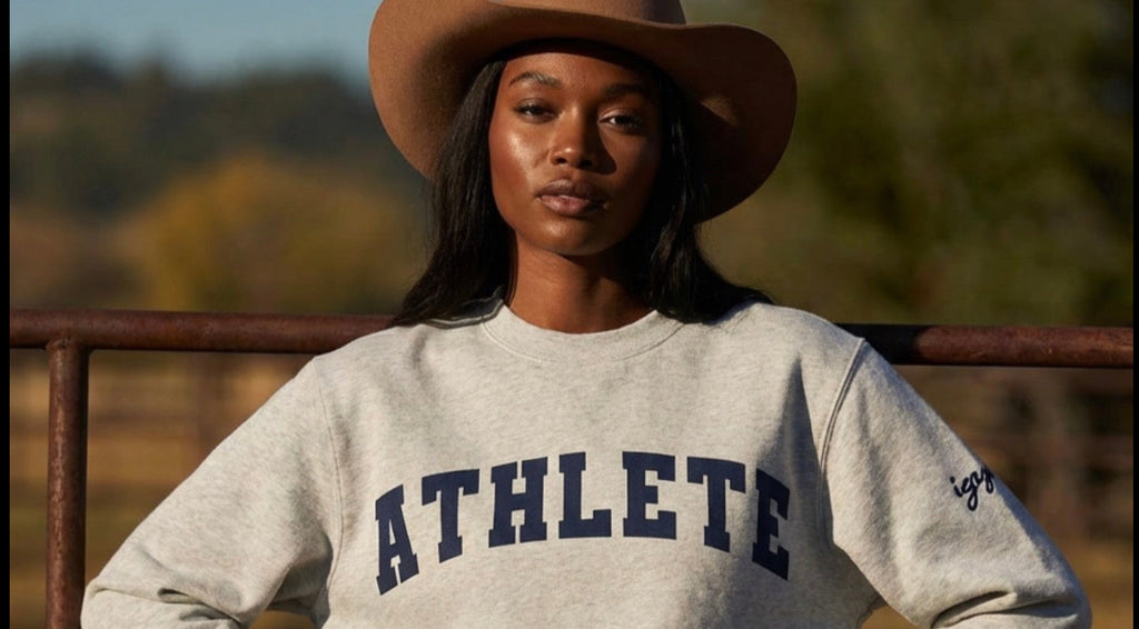 Person wearing a gray sweatshirt with 'ATHLETE' printed on it, standing in front of a rustic metal gate.